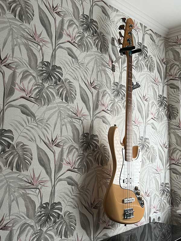 S8_600 Beige E-Bass-Gitarre, die an einer Wand mit tropisch gemusterter Tapete in gedeckten Farben hängt, kombiniert mit einer schlichten weißen Zierleiste an der Decke.